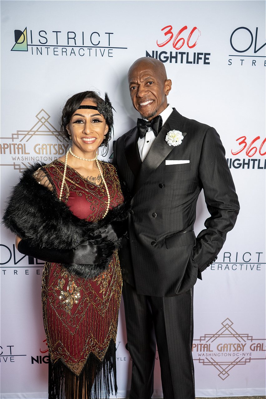 A couple in Great Gatsby theme clothing at Capital Gatsby New Year's Eve DC Gala at HQO pose in front of the step and repeat.